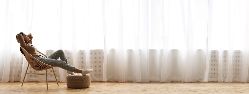 Woman sitting in front of a large window with sheer drapes relaxing in a chair with feet propped up and arms behind her head | Whole-Home Air Purifier Arlington and Mansfield, TX